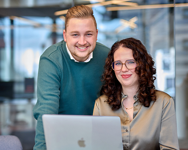 Mann stehend leicht nach vorne gebeugt, Frau sitzend vor Laptop, beide mit freundlichem Blick in die Kamera